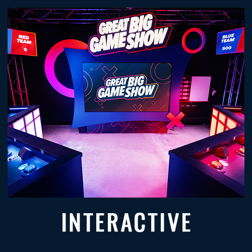 Colorful game show stage with buzzers and screens for red and blue teams, labeled “INTERACTIVE”