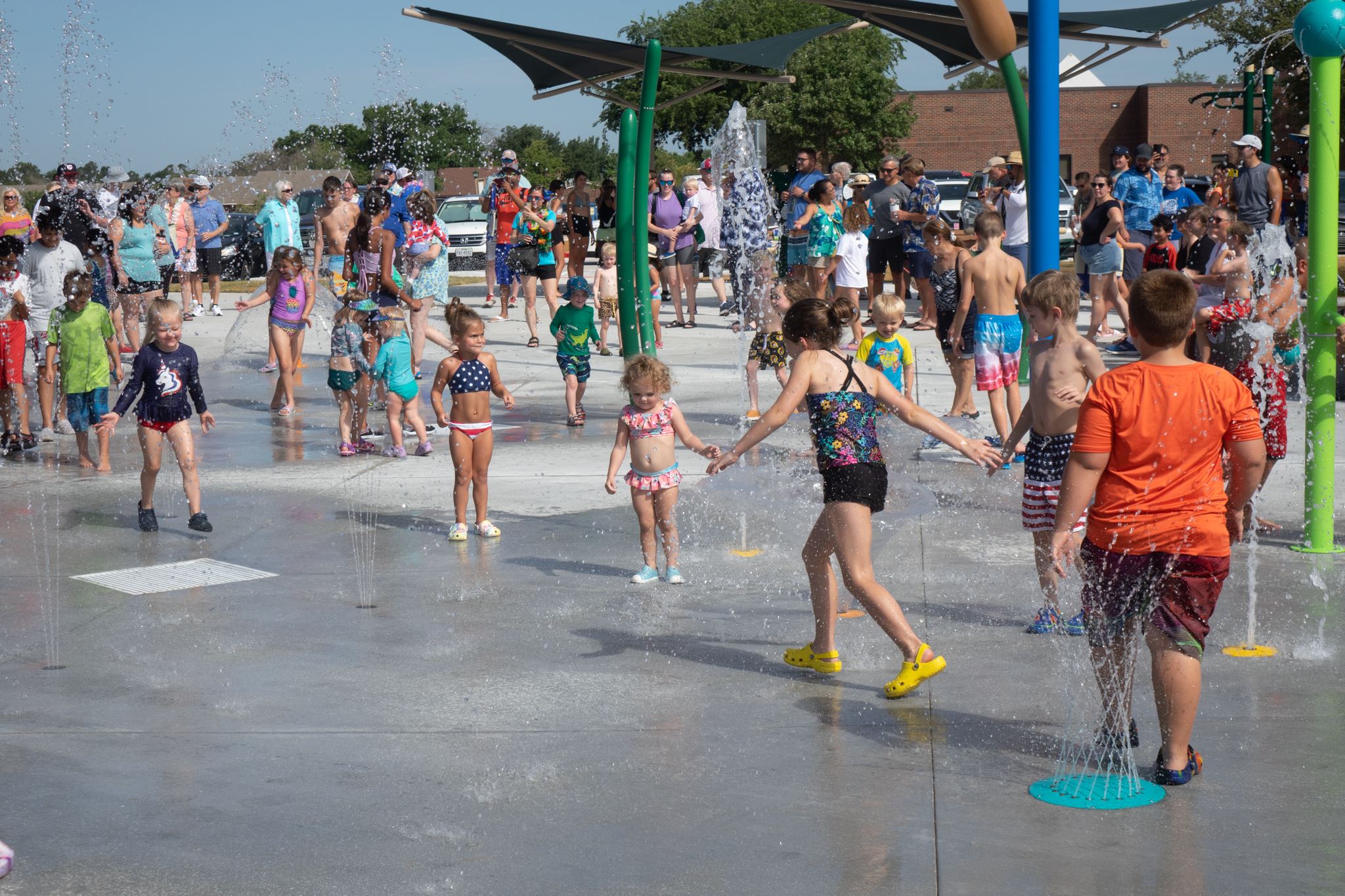 Kids playing in fountains at a crowded splash pad on a sunny day, with families watching nearby.